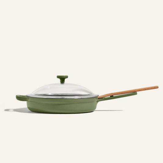 The Cast Iron Always Pan, featuring a green enamel finish, a wooden handle, and a glass lid, is pictured against a plain light background.