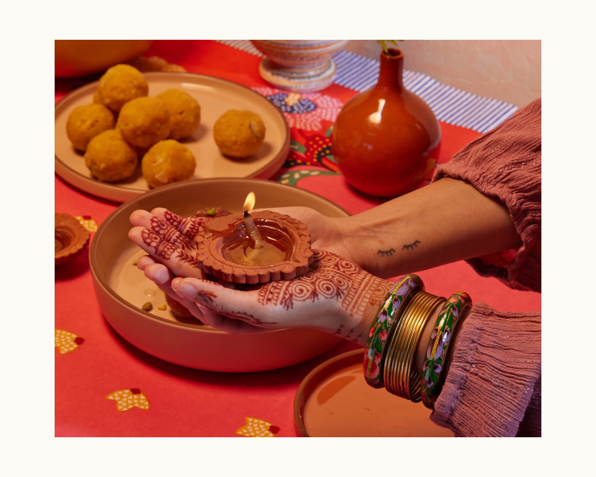 During the Diwali celebration, a person with henna and colorful bangles holds a lit clay lamp over a table displaying sweets, festive decor, and the Golden Fry Set on a cooling rack.