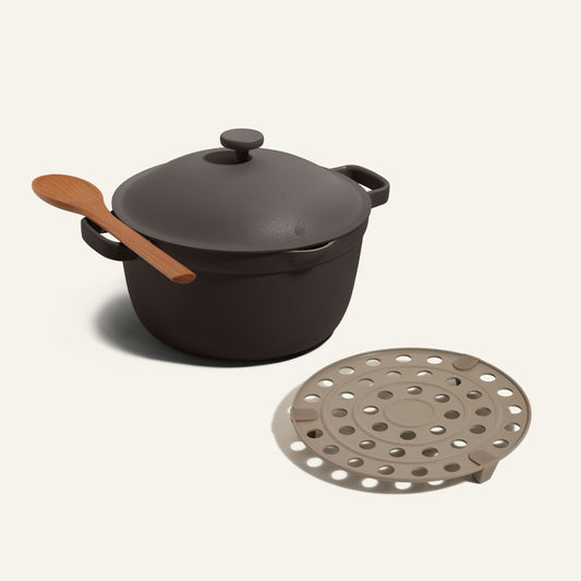 A Perfect Pot with lid and Roasting Rack, featuring a wooden spoon on the handle, sits beside a round, beige perforated steamer insert.