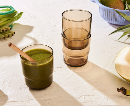 A glass of green smoothie with a wooden spoon sits beside two stacked Short Night + Day Glasses on a textured white surface, with fruit and a blue basket in the background.