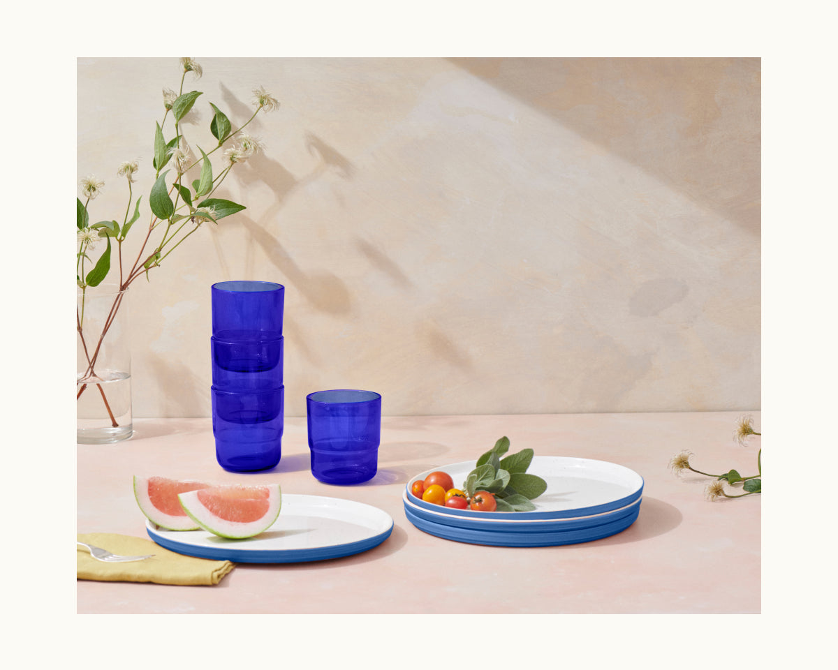 A set of blue-rimmed white plates with tomatoes and watermelon, colorful Short Night + Day Glasses stacked, a glass vase with branches, and a yellow napkin on a light pink surface.