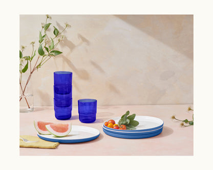 A set of blue-rimmed white plates with tomatoes and watermelon, colorful Short Night + Day Glasses stacked, a glass vase with branches, and a yellow napkin on a light pink surface.