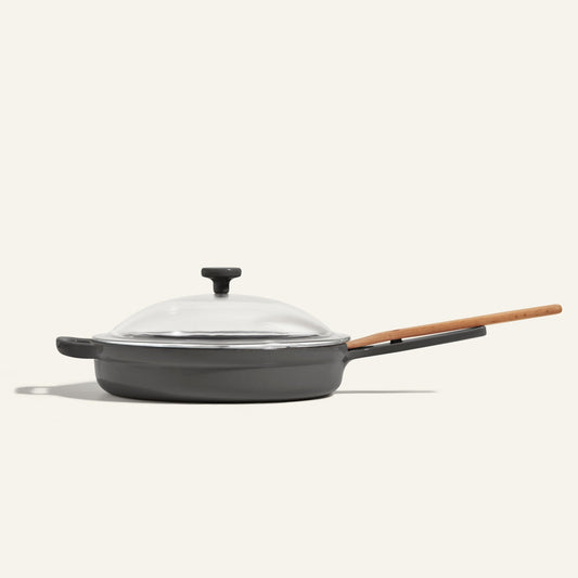 The Cast Iron Always Pan, finished in black enamel with a wooden handle and transparent glass lid, is displayed against a plain light background.