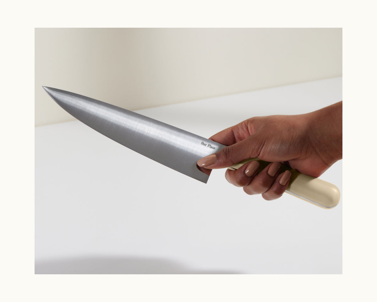 A hand holds the Everyday Chef’s Knife, featuring a full-tang German steel blade and a light-colored handle, poised over a white surface.