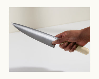 A hand holds the Everyday Chef’s Knife, featuring a full-tang German steel blade and a light-colored handle, poised over a white surface.