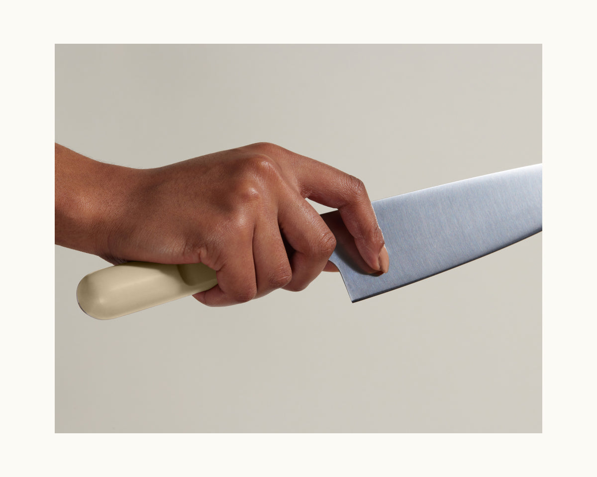 A hand grips the full-tang blade of the Everyday Chef’s Knife with a beige handle near the bolster, shown against a plain light background.