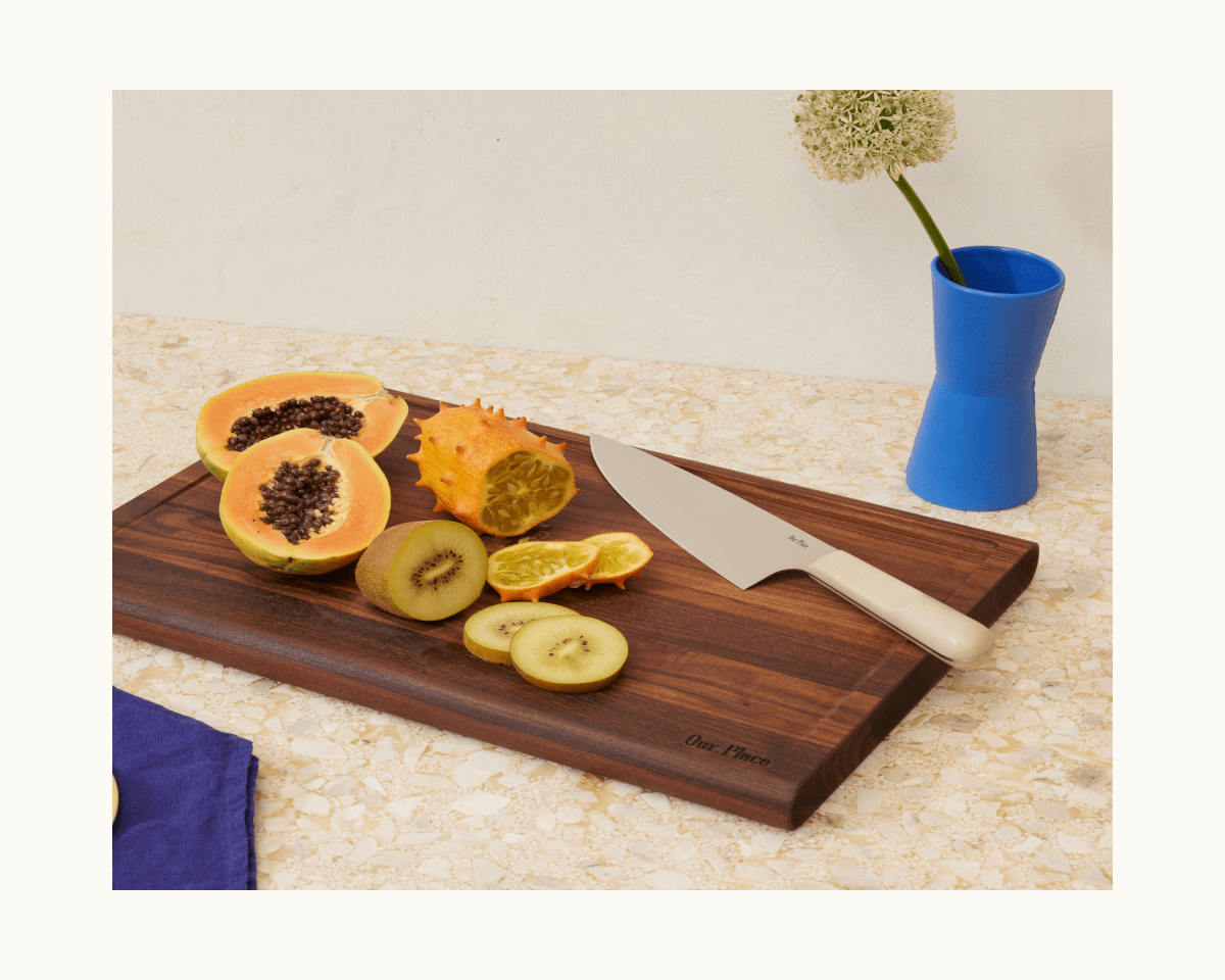 A cutting board with sliced papaya, kiwano, kiwi, and orange sits beside the Everyday Chef’s Knife; a blue vase with a white flower brightens the background.