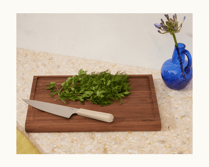 The Everyday Chef’s Knife rests beside fresh herbs on a wooden cutting board atop a speckled countertop, with a blue vase of purple flowers nearby.