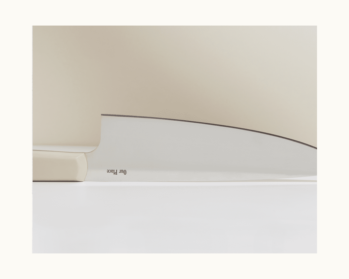 The Everyday Chef’s Knife, featuring a white handle and a partially visible German steel blade, rests on a white surface with a beige background.