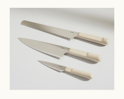 The Everyday Chef’s Knife, featuring a beige handle and crafted from durable German steel, is displayed on a white surface alongside a bread knife and paring knife.