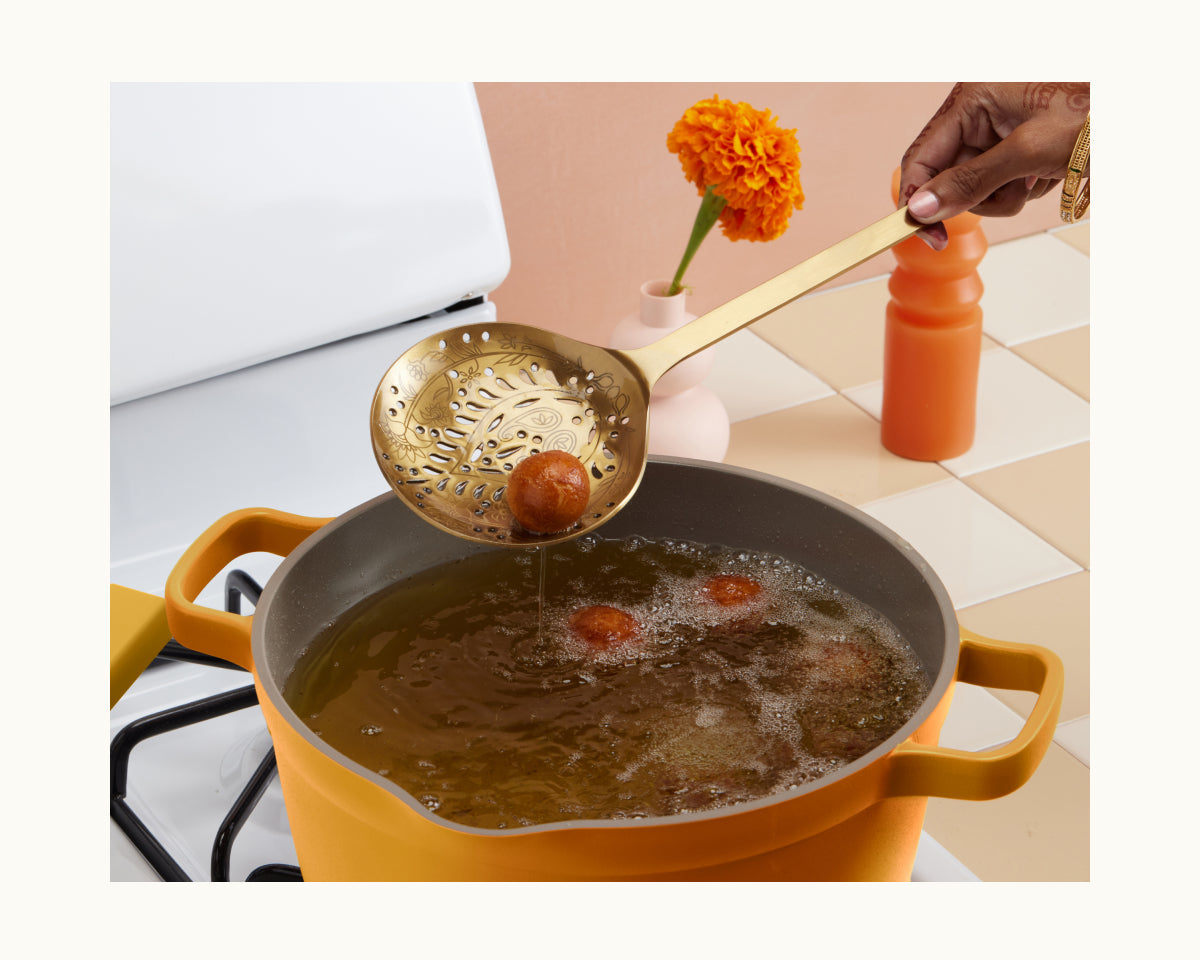 Using the Golden Fry Set, a person lifts a fried dessert ball from hot oil in a yellow pot, with marigold flowers and a bottle nearby, evoking the festive spirit of Diwali.