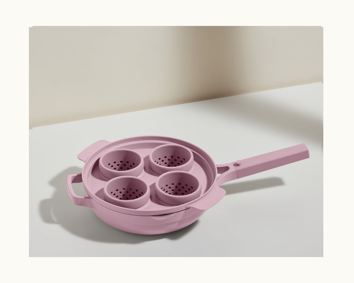 The Egg Poacher is a pink multi-purpose saucepan, similar to the Always Pan, with four round silicone cups for poaching eggs. It’s displayed elegantly on a light-colored surface.
