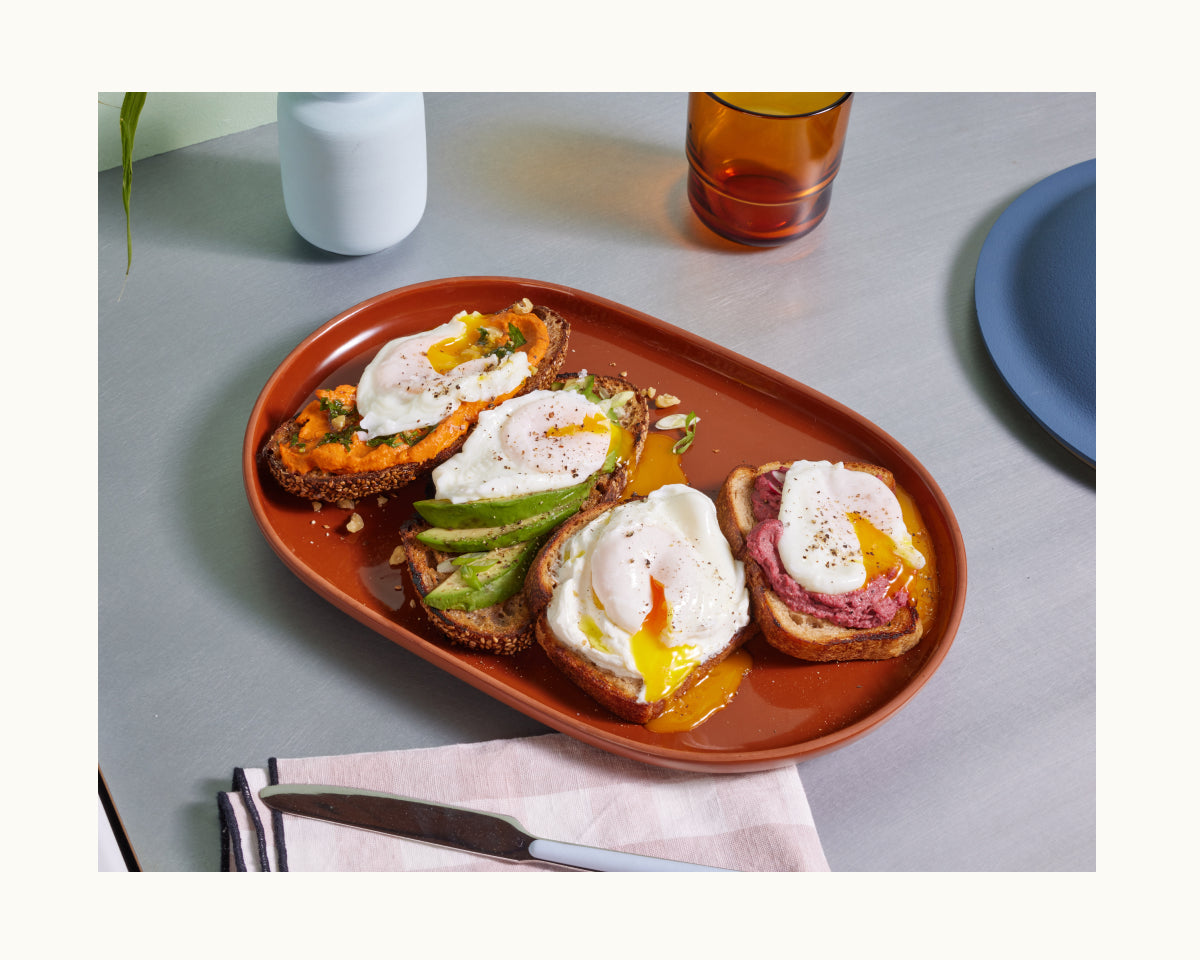 A brown oval plate with four slices of toast topped with poached eggs made using the Egg Poacher and various spreads sits on a table with a knife, napkin, glass, and an Always Pan nearby.