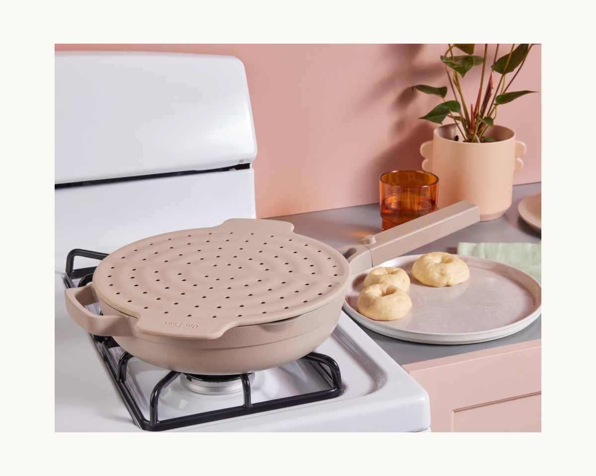 A beige Fearless Fry with a perforated steamer insert rests on the stove next to three dough balls, while a matching utensil holder and plant decorate the background, highlighting chic splatter prevention in your kitchen.