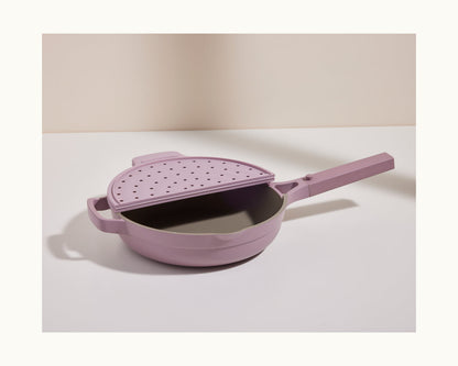 A lavender Fearless Fry pan with a matching strainer lid and long handle sits on a white surface, providing stylish splatter prevention against a neutral backdrop.