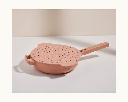 The Fearless Fry, a pink nonstick frying pan inspired by the Always Pan, comes with a matching handled lid featuring multiple steam holes for splatter prevention and sits on a white surface against a neutral background.