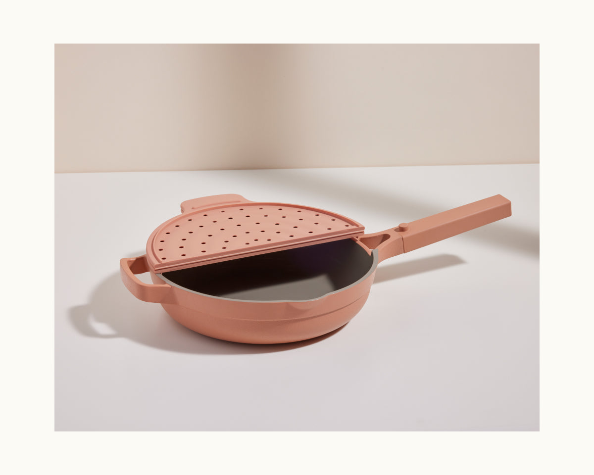 The Fearless Fry is a pink nonstick frying pan featuring a matching handle and a fitted perforated lid for splatter prevention, displayed on a white surface with a neutral background.