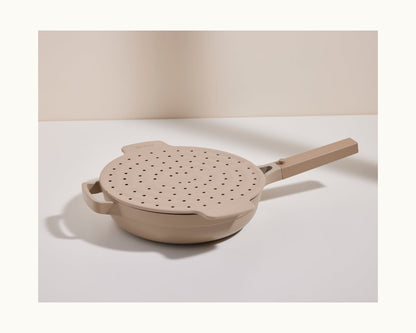 A beige Fearless Fry pan with a long handle and a perforated splatter-prevention lid rests on a white surface against a neutral background.