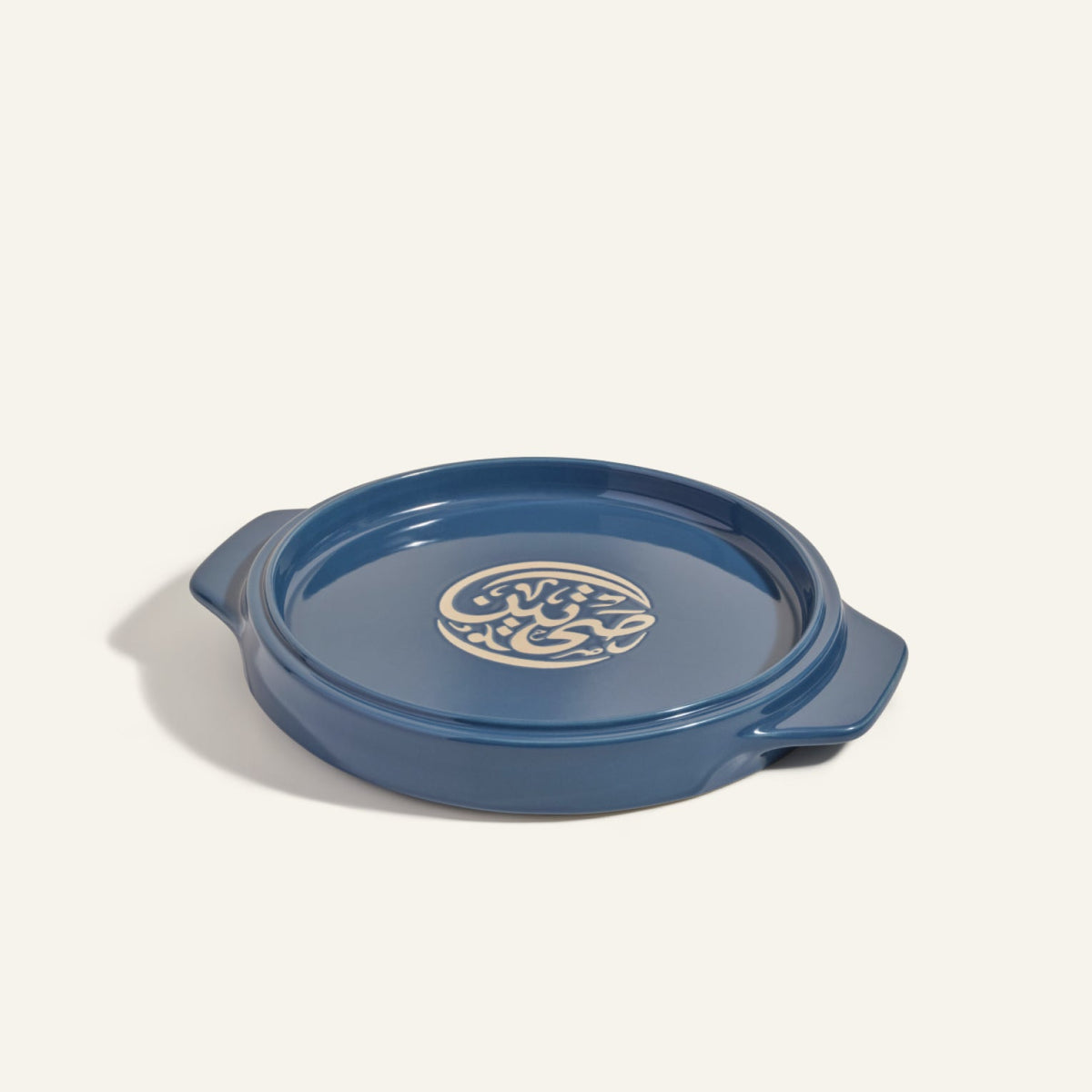 The Flipping Platter is a round blue ceramic server with side handles and a cream abstract lid design, ideal for serving dishes like tahdig or maqluba. Shown on a light background.
