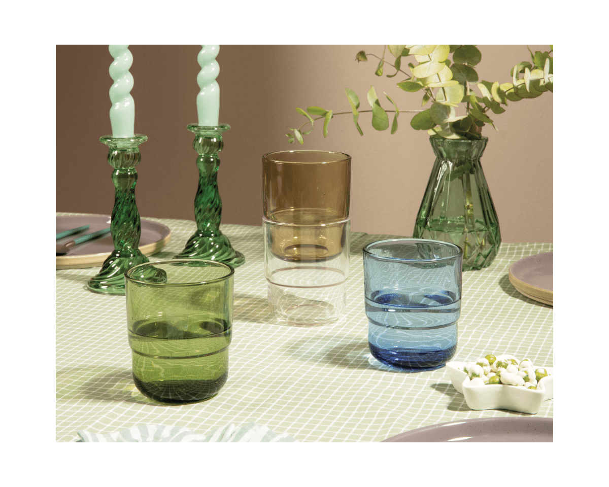 Three Short Night + Day Glasses in green, brown, and blue sit on a checkered table beside green candlesticks, candles, a vase with foliage, and a small dish.