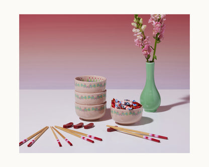 The Rice Bowl Set includes four pink stoneware bowls, stacked beside a green vase with pink flowers and four pairs of chopsticks with heart-shaped rests, displayed on a white surface.