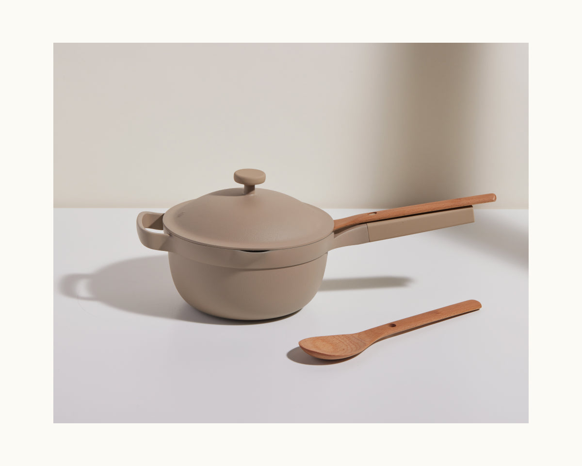 A beige saucepan with a lid and long handle sits on a white surface beside Beechwood Spoons, making an ideal addition to your collection of kitchen utensils.