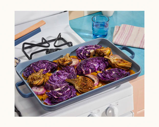 A baking tray from a Bakeware Set holds purple cabbage, artichokes, onions, and yellow vegetables on a stovetop next to a glass of water and a folded cloth.