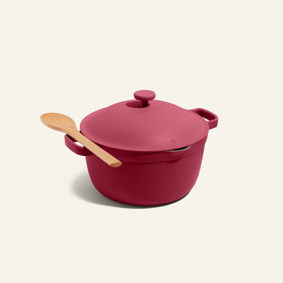 The Perfect Pot in red comes with a lid and features a wooden spoon resting on its handle, showcased against a plain light background.
