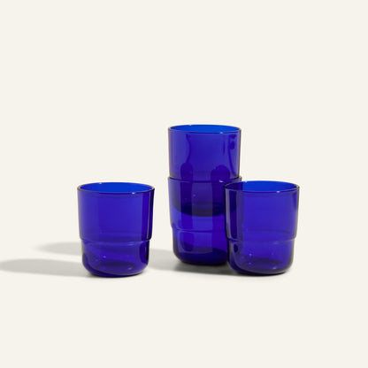 Four Short Night + Day Glasses in cobalt blue recycled glass are stacked on a neutral background, highlighting vibrant drinking tumblers ideal for any table setting.