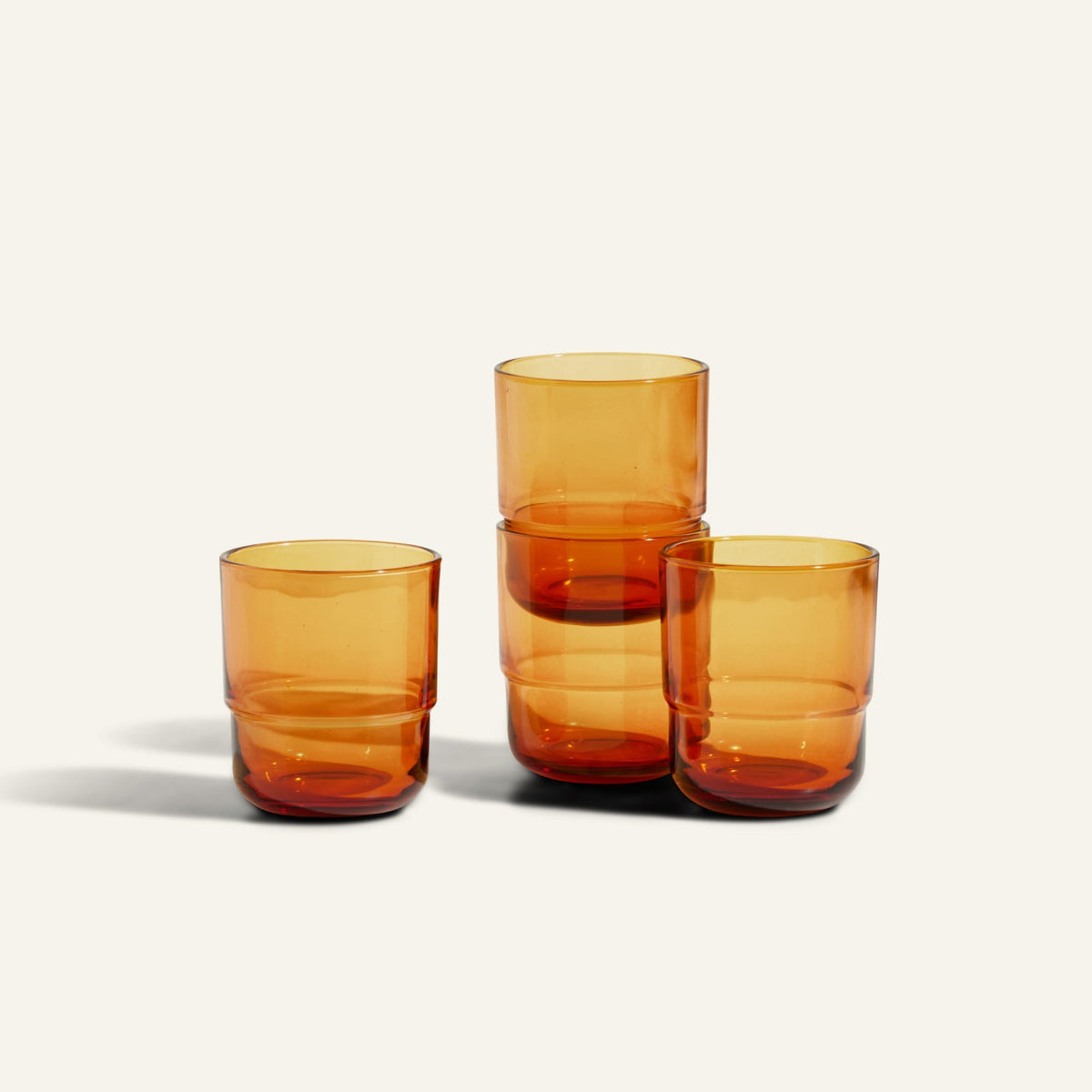 Four Short Night + Day Glasses in amber recycled glass are arranged on a light surface, with two stacked on the right and two placed side by side on the left.