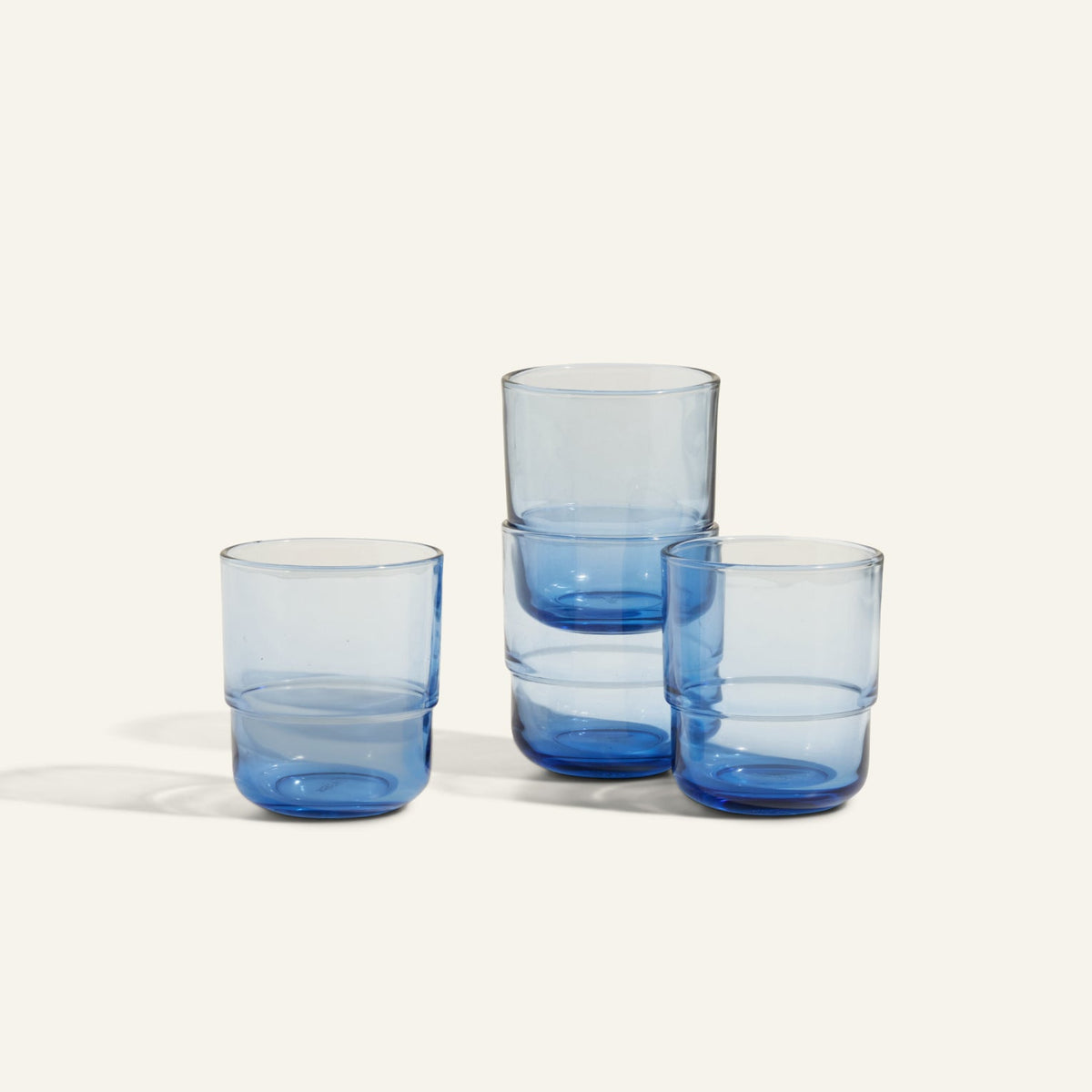 Four Short Night + Day Glasses made from blue recycled glass are stacked in pairs on a plain, off-white background.
