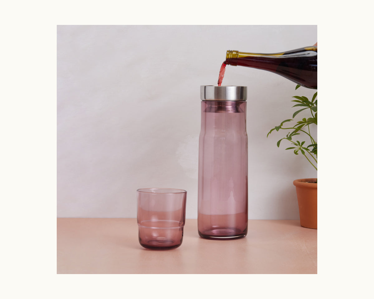 A dark liquid is poured from a bottle into the Night + Day Carafe, which stands next to a matching glass cup and a potted plant on a light surface.