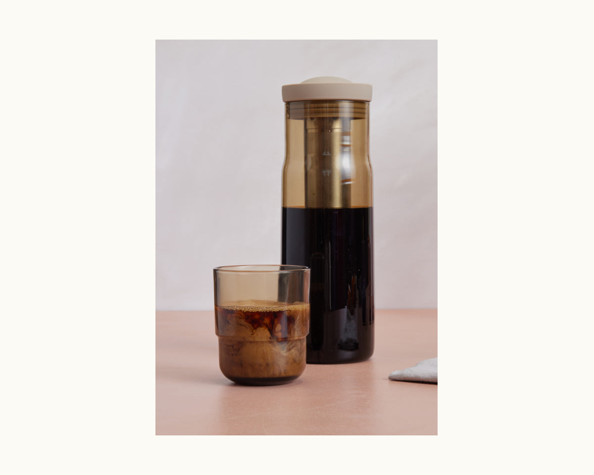 A glass of iced coffee with cream sits beside the Night + Day Carafe cold brew infuser on a light pink surface.