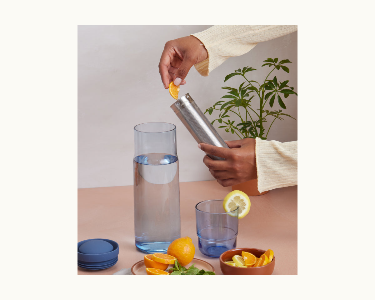 Someone drops a lemon slice into the Night + Day Carafe filled with water beside a glass with a lemon slice, blue lids, fresh lemons, mint, and a potted plant in the background.