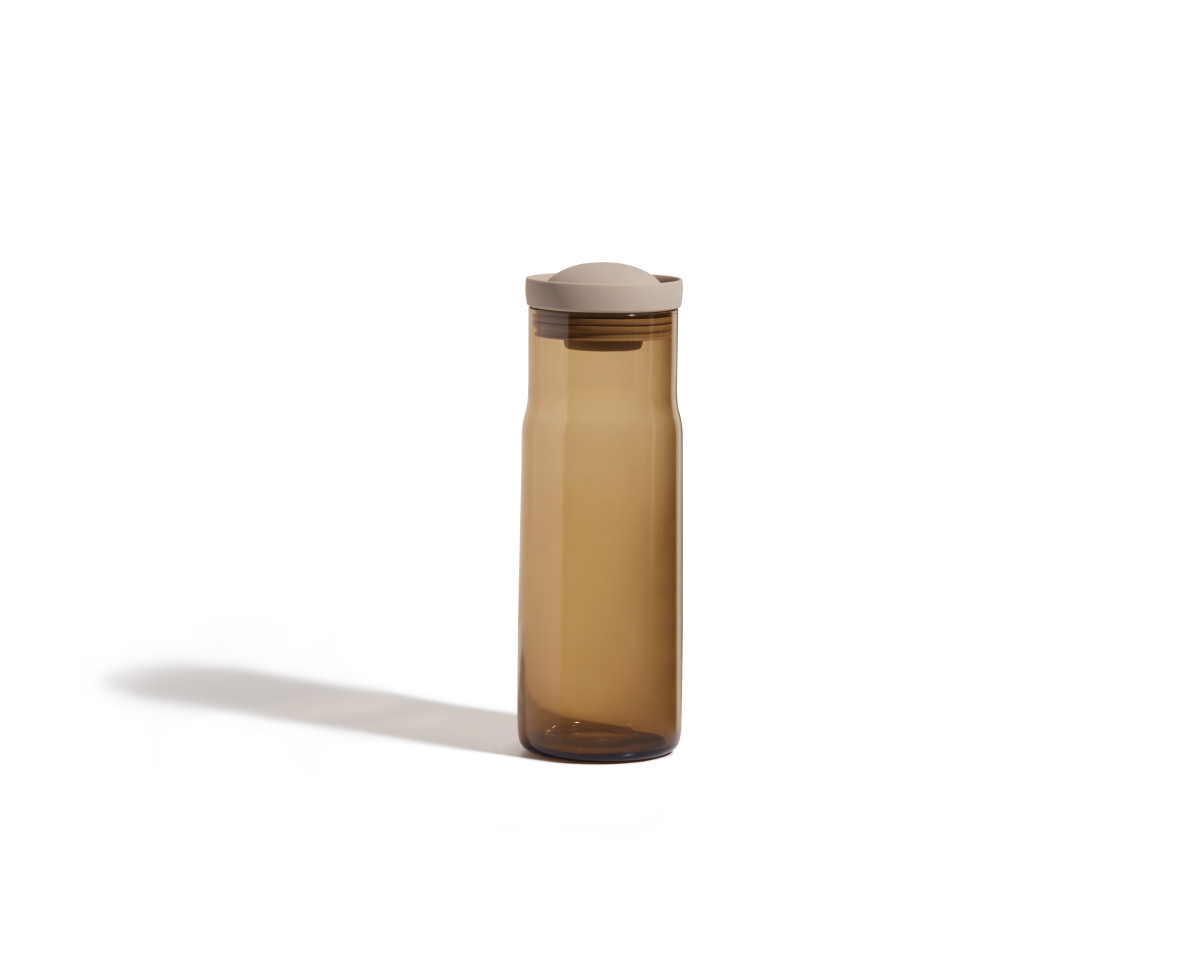 A translucent brown Night + Day Carafe with a beige lid stands upright on a plain white background, its tall sleek shape resembling a modern water bottle.