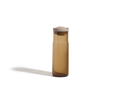 A translucent brown Night + Day Carafe with a beige lid stands upright on a plain white background, its tall sleek shape resembling a modern water bottle.