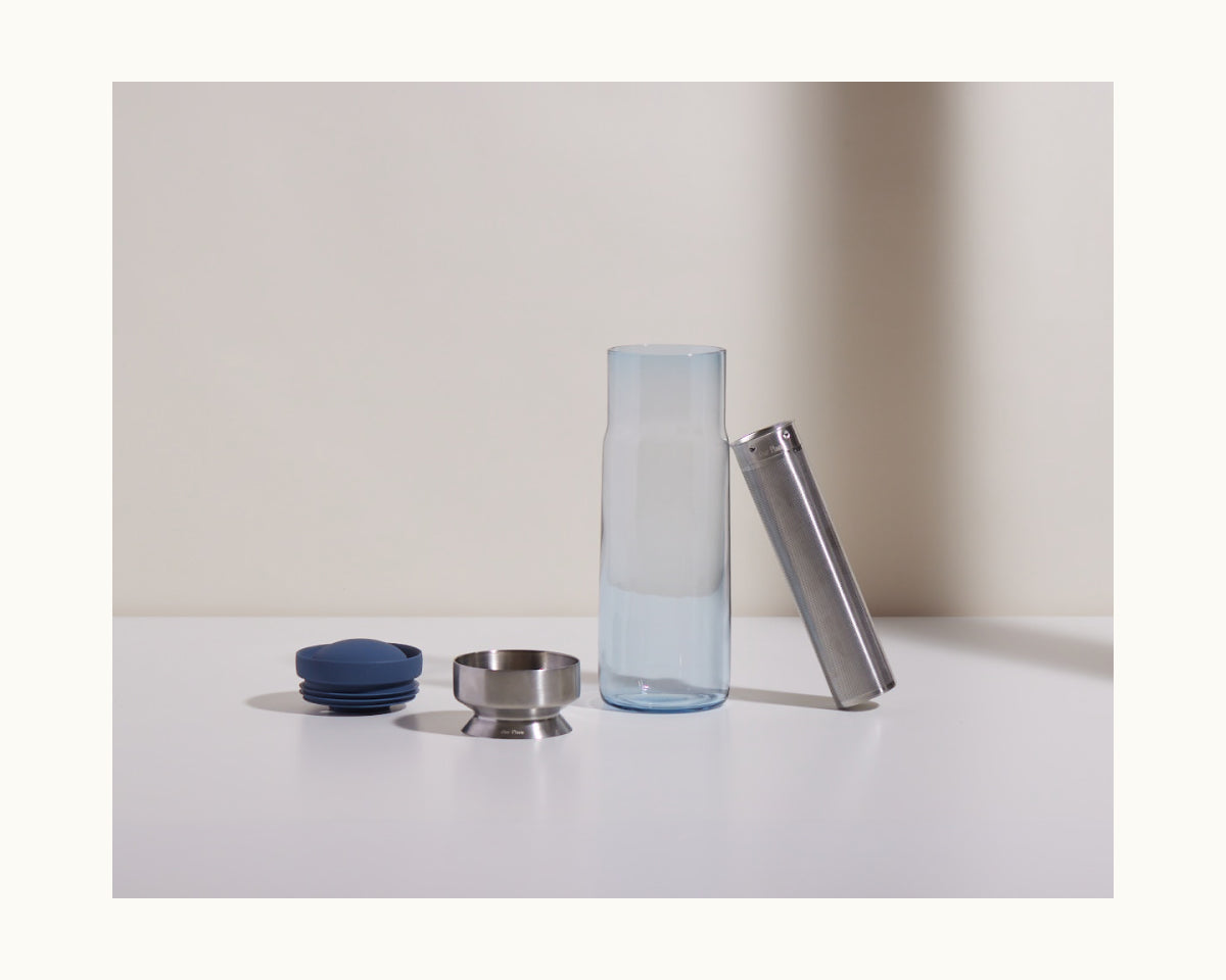 The Night + Day Carafe is shown disassembled into four parts—a blue cap, metal ring, glass body, and cylindrical metal filter—arranged neatly on a white surface, perfect for cold brew infusing.