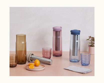 The Night + Day Carafe, with its metal infuser and colored lid, is displayed on a table alongside glasses, sliced citrus fruits on a plate, a cloth napkin, and a small potted plant.