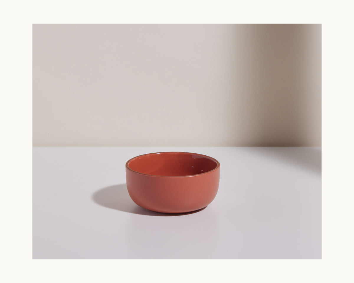 A small, round, terracotta-colored Soup Bowl from the Soup Bowls set rests on a white surface against a plain, light backdrop.