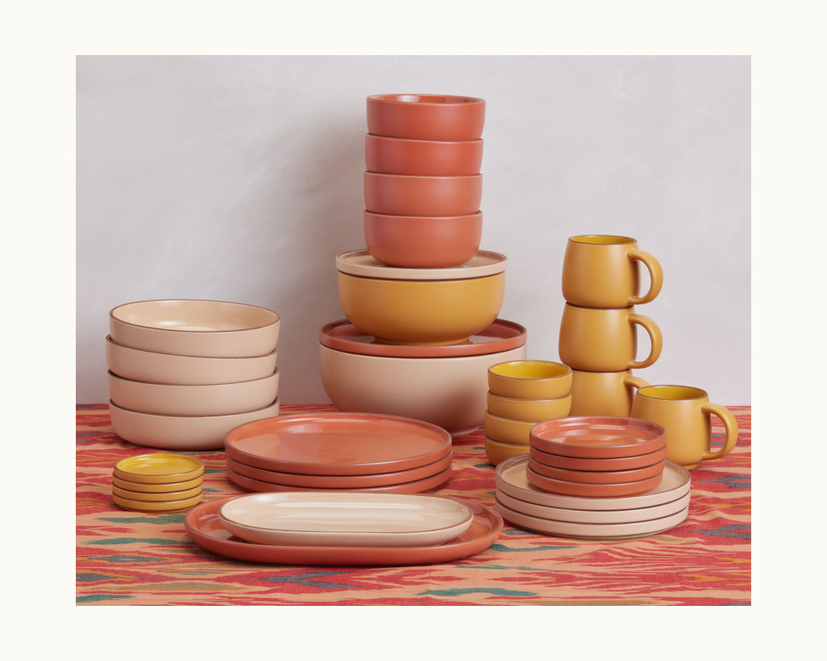 The Soup Bowls set features stackable bowls, plates, and mugs in earthy beige, terracotta, and mustard tones, arranged stylishly on a patterned tablecloth.