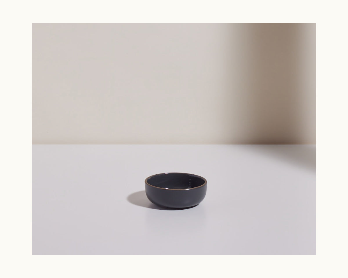 A Tiny Bowls black ceramic piece sits on a white surface with a right-side shadow against a neutral background—ideal for modern kitchens wanting stylish, stackable tableware.