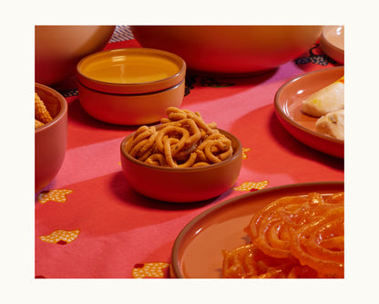 A bowl of crunchy Indian snacks sits on a red tablecloth among ceramic bowls and plates, highlighting Tiny Bowls—stylish, stackable tableware perfect for festive gatherings.