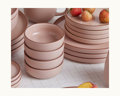 A stack of four pink Tiny Bowls, which are scratch resistant and ceramic, sits next to matching plates, mugs, and several strawberries on a white checkered surface.