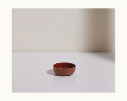 A Tiny Bowls ceramic piece, small and brown, sits empty on a white surface before a beige wall; its design is stackable for versatile tableware use.