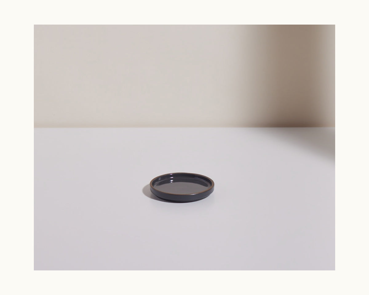 The Tiny Plates product is a small, round black ceramic plate placed on a white surface with a light background, casting a shadow to the right.