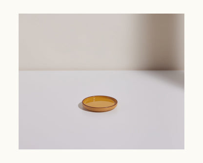 The Tiny Plates are shallow, round ceramic plates with an amber hue, displayed on a white surface against a plain, light background.