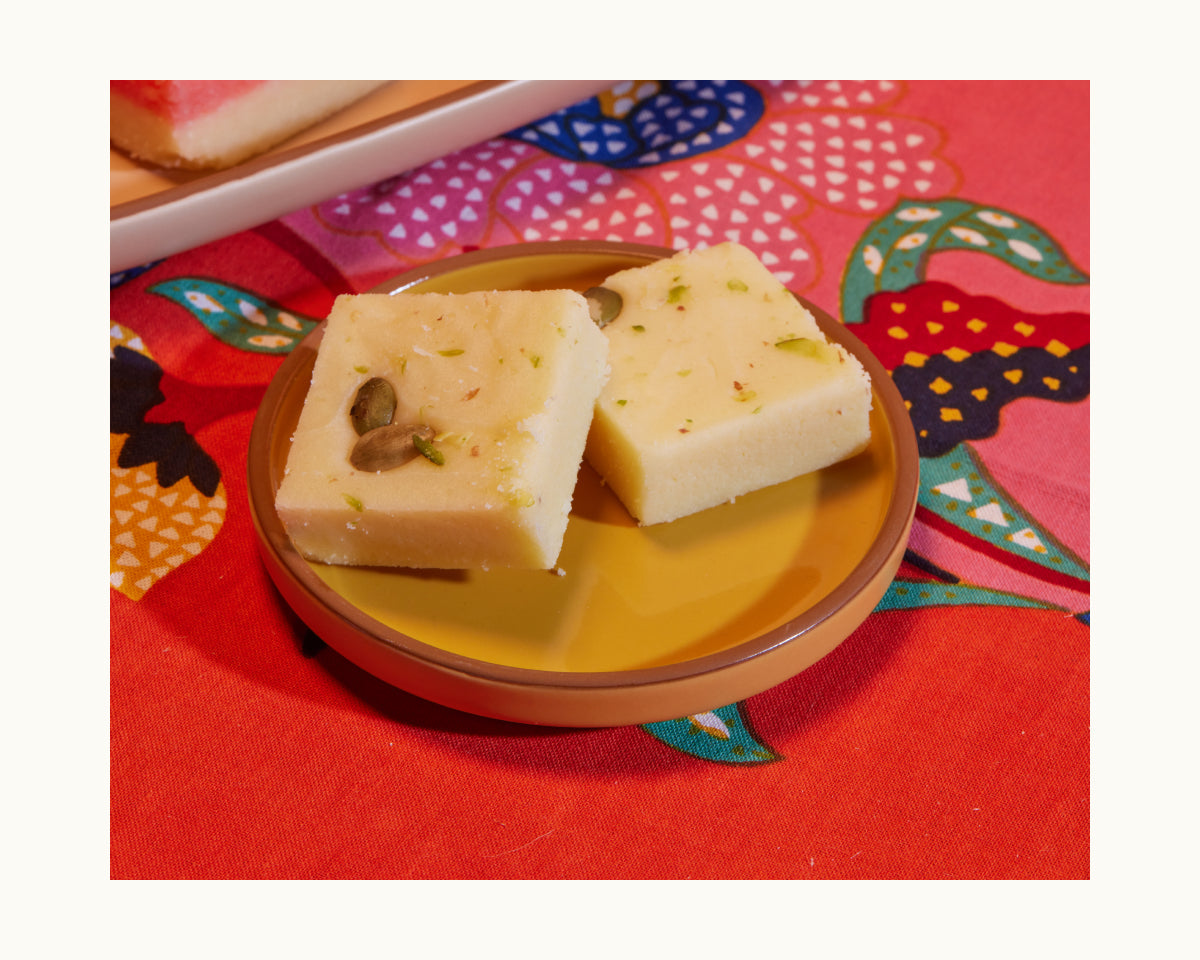 Two pieces of white Indian sweet topped with nuts are served on Tiny Plates, a set of brown ceramic snack plates, displayed on a colorful patterned cloth.