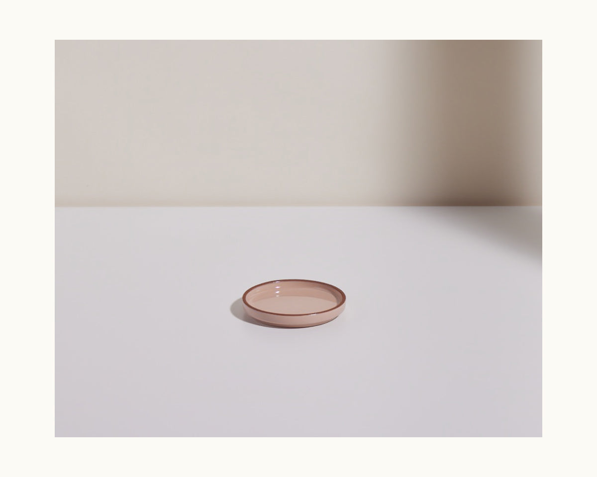 A Tiny Plates snack plate, small and round in pink, rests on a white surface with a soft shadow against a neutral background.