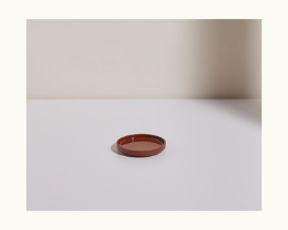A Tiny Plates small, round brown ceramic plate rests on a white surface with a pale backdrop and a shadow cast to the right.