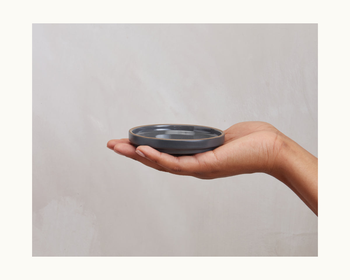 A person’s hand holds a round, flat, dark gray Tiny Plates ceramic plate from a tableware set against a plain light background.